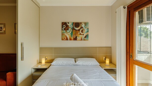 Una habitación de hotel con una cama, lámparas de noche, un cuadro en la pared y vistas a través de un ventanal.