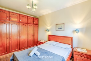 Une chambre à coucher avec une armoire en bois, un lit recouvert de draps blancs, une couverture bleue et une table de chevet avec une lampe.