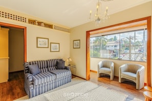 Un salon comprenant un canapé, deux fauteuils, un lustre et une grande fenêtre donnant sur des maisons.