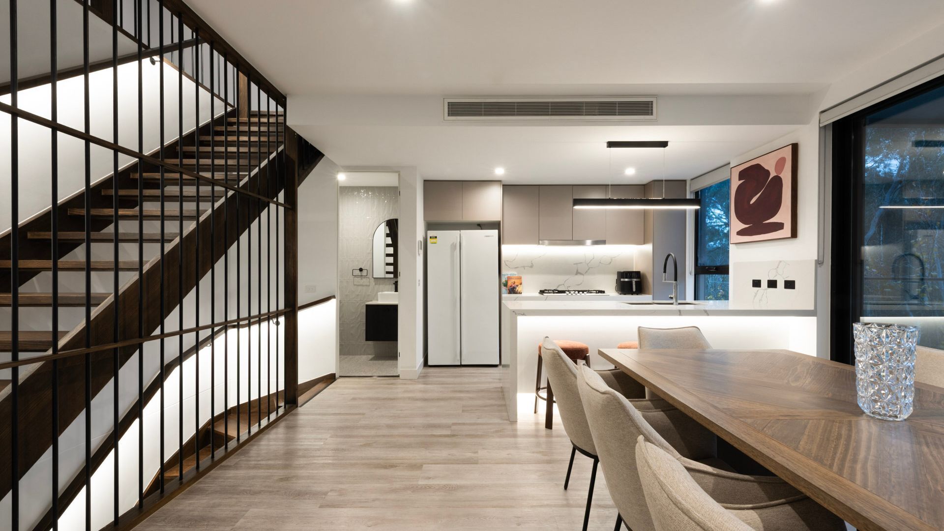 Eine moderne Küche mit einer zentralen Kochinsel, ein Essbereich mit einem Holztisch und eine Treppe mit Metallgeländer.