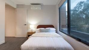 Um quarto com cama, luminária de cabeceira, janela e aparelho de ar condicionado.