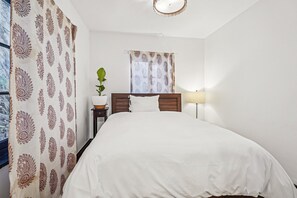 Ein Schlafzimmer mit Bett, Nachttisch, Lampe, einem Fenster mit Vorhängen und einer Topfpflanze.
