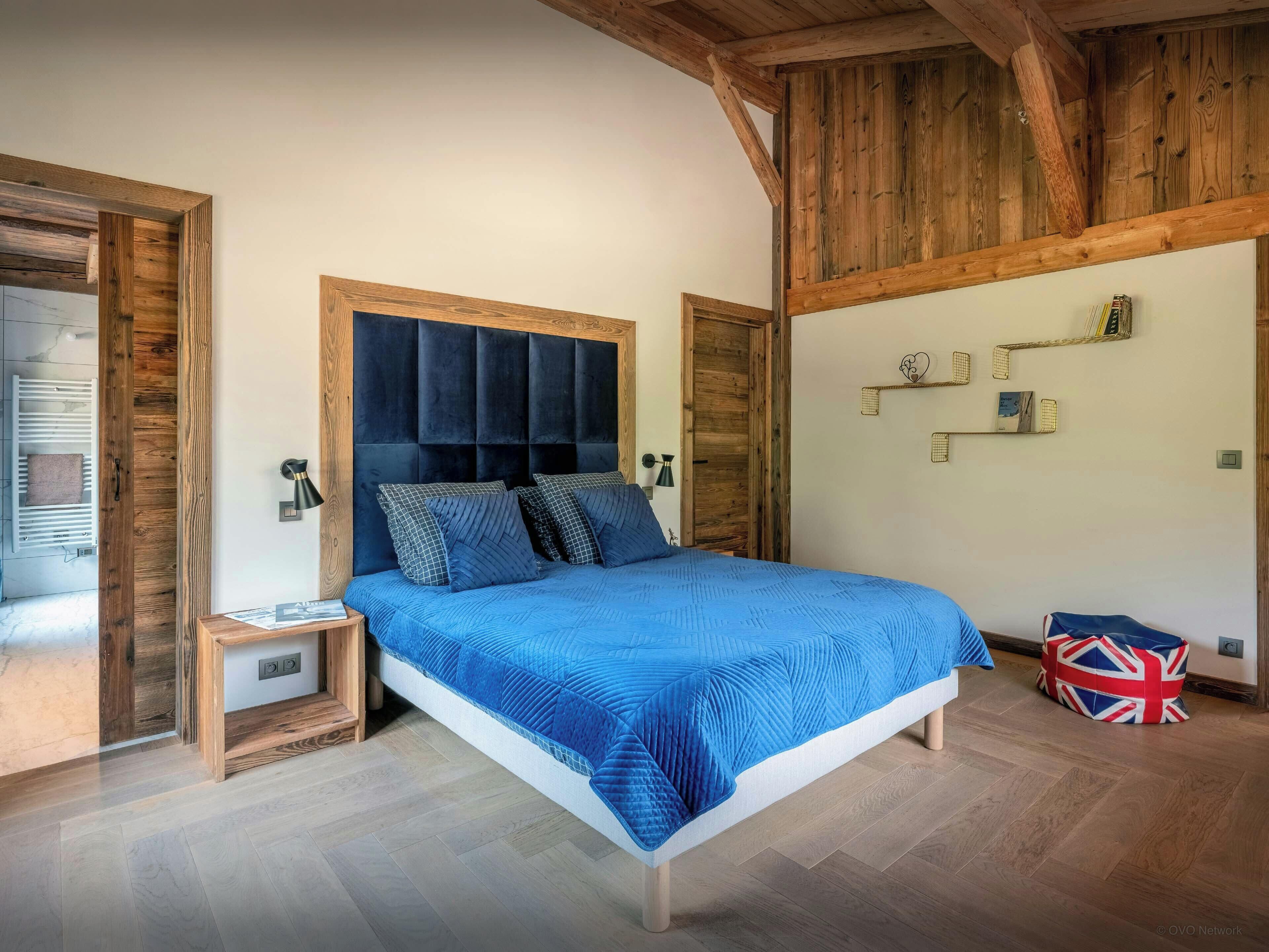 A bedroom with a wooden headboard, a blue bedspread, bedside lamps, and a small bedside table.