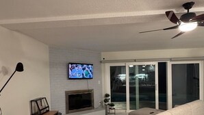 Un salon comprenant un canapé, un téléviseur fixé au mur, une cheminée, un ventilateur de plafond et un coin repas avec une table et des chaises.