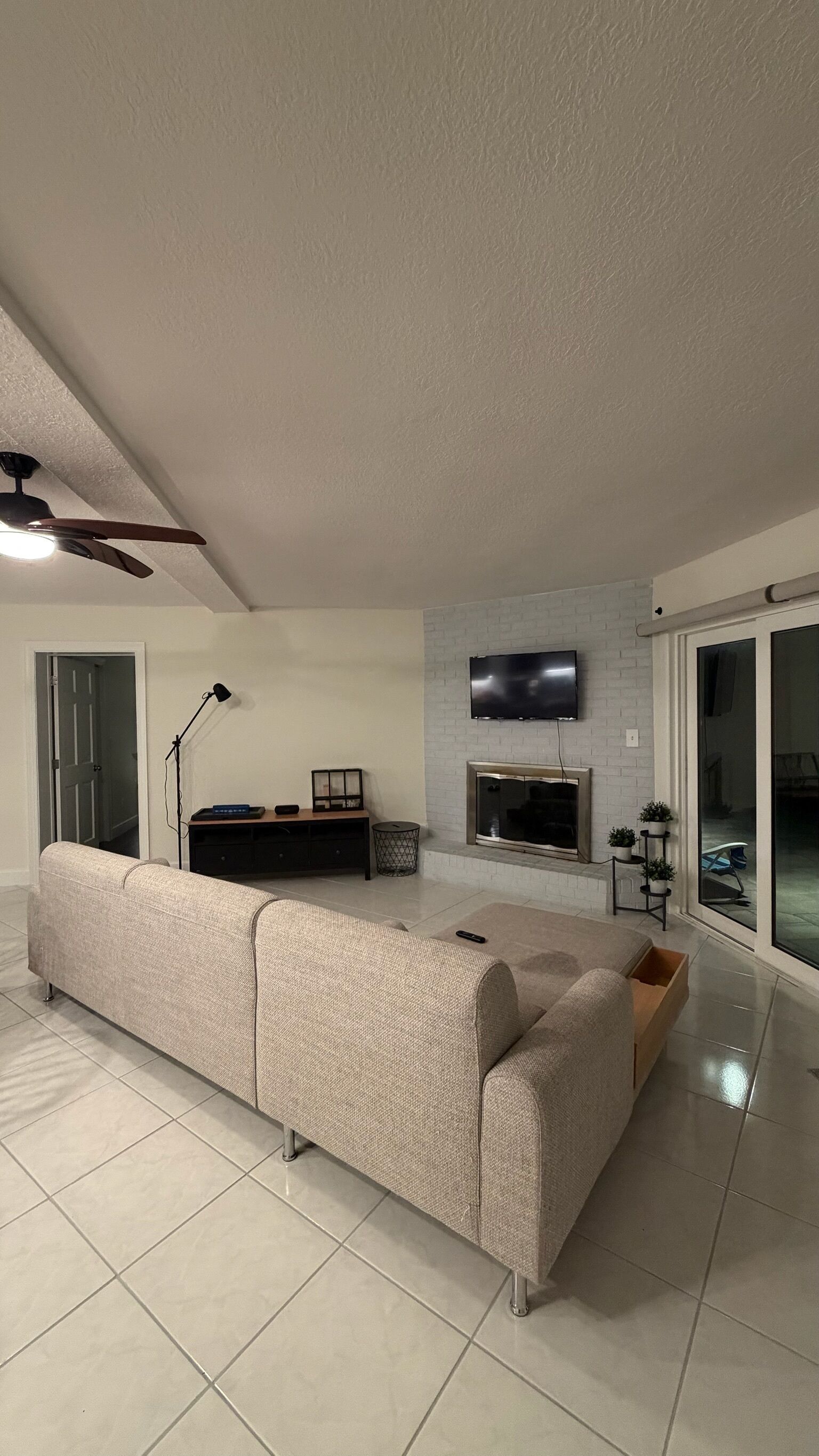 A modern living room with a large sofa, a flat-screen TV mounted on a brick wall, and a ceiling fan.