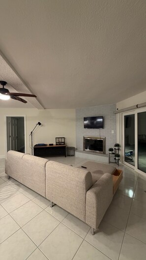A modern living room with a large sofa, a flat-screen TV mounted on a brick wall, and a ceiling fan. - Beach House in Indian Rocks Beach | 4 bedrooms with pool (Indian Rocks Beach)