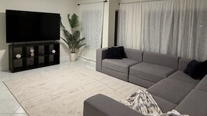 Un salon avec un téléviseur à écran plat, un canapé gris, un plaid à motifs et une plante en pot.