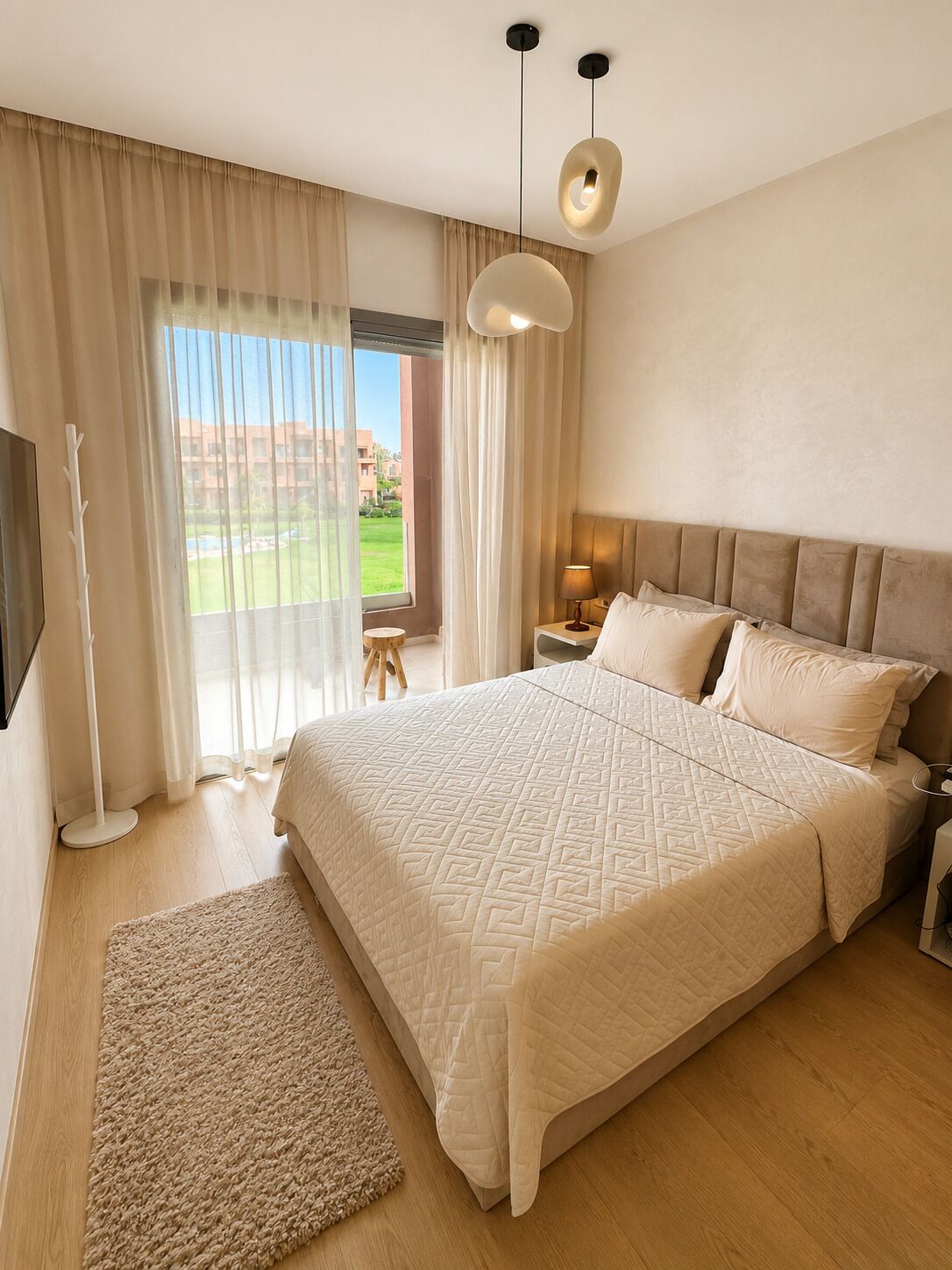 A hotel room with a large bed, a bedside table with a lamp, a floor lamp, and a view of a building and greenery outside.