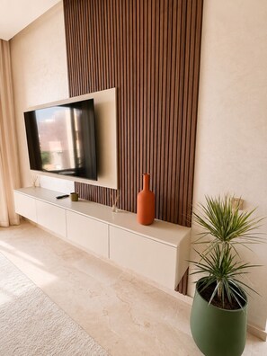 A modern living room with a flat-screen TV mounted on a wall, a white console table, a potted plant, and a large vase. - 2-bedroom apartment, 2 pools, Golf Prestigia Jubaea (Marrakesh)