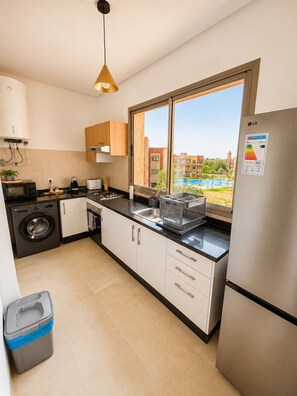 A modern kitchen with a built-in washing machine, a stainless steel refrigerator, and a window with a view of a swimming pool and buildings. - 2-bedroom apartment, 2 pools, Golf Prestigia Jubaea (Marrakesh)