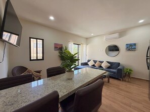 A modern living room with a dining area, a sofa set, a potted plant, and a flat-screen TV mounted on the wall. - Modern Condo in Gabbiani, by GPS (Mazatlán)