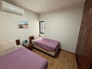 A bedroom with two beds, wooden flooring, a wardrobe, a window, and an air conditioner. - Modern Condo in Gabbiani, by GPS (Mazatlán)