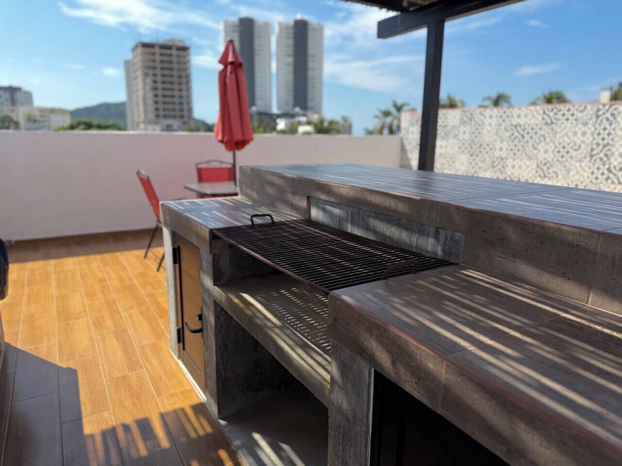 An outdoor patio with a grill, a red umbrella, and a view of city buildings.