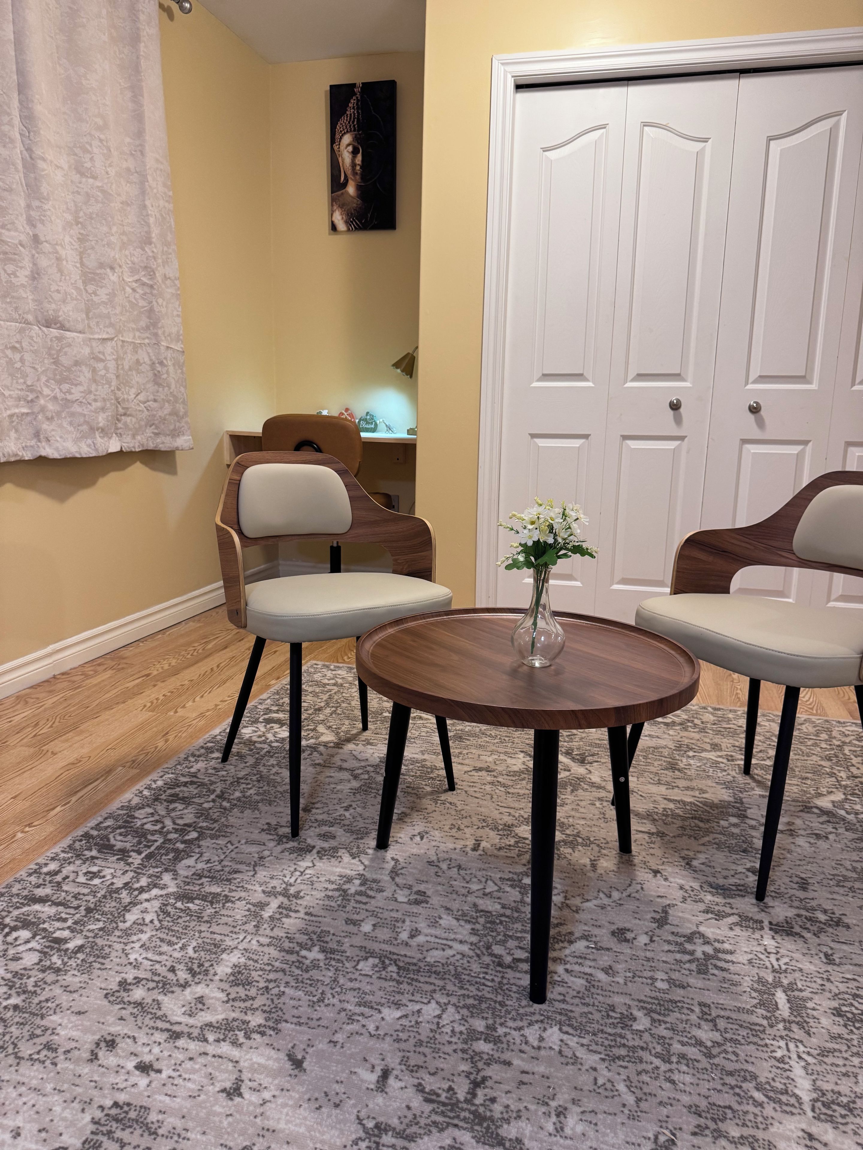 Um quarto com uma mesa de madeira redonda, duas cadeiras, um vaso com flores, uma pintura de Buda e uma porta branca.