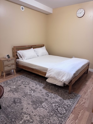Een eenpersoonsbed met houten frame en witte beddengoed, een nachtkastje met een lamp, en een wandklok.