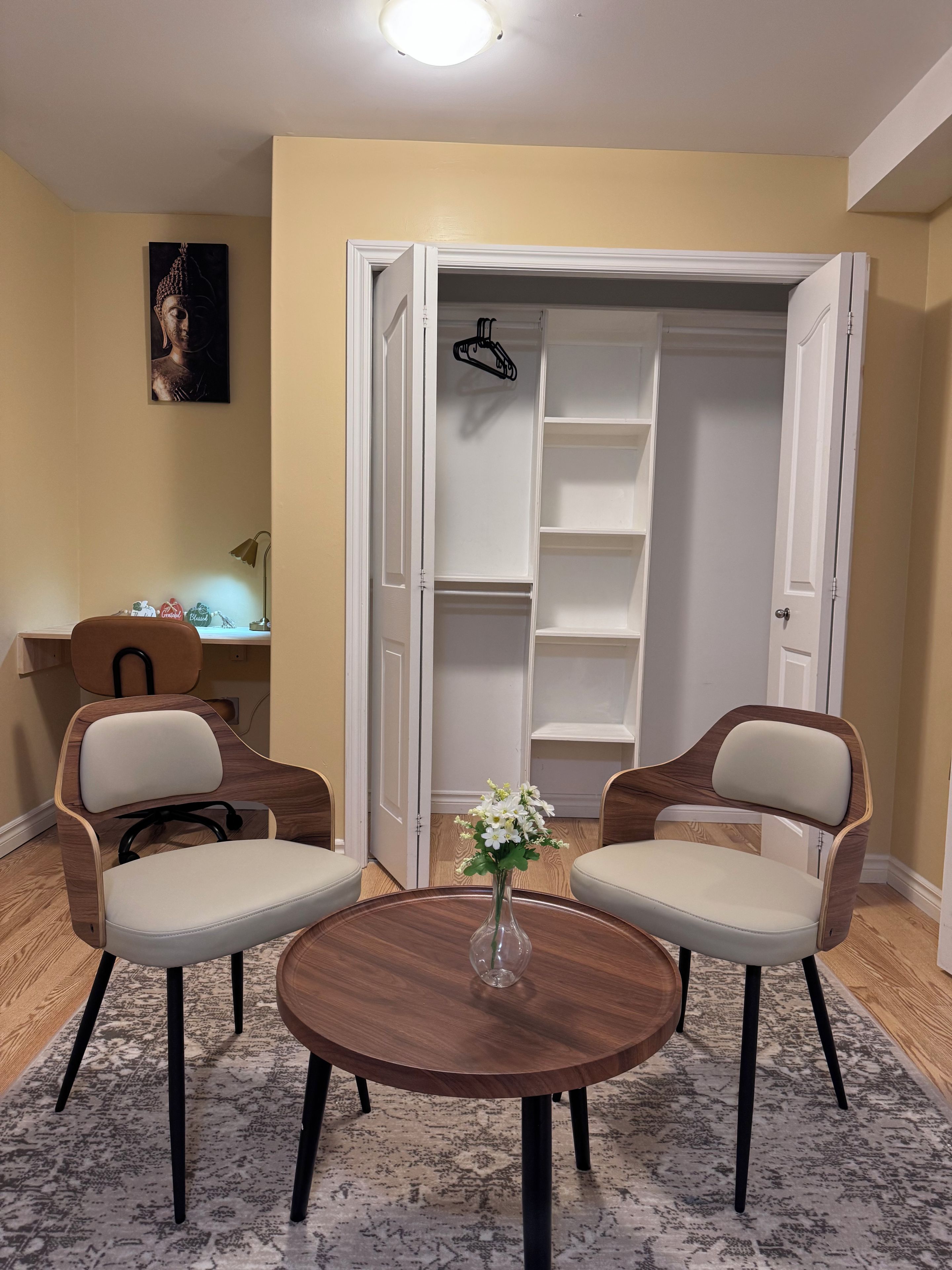 Une pièce comprenant deux chaises, une petite table, un vase avec des fleurs, un bureau avec une lampe et une armoire avec des étagères.