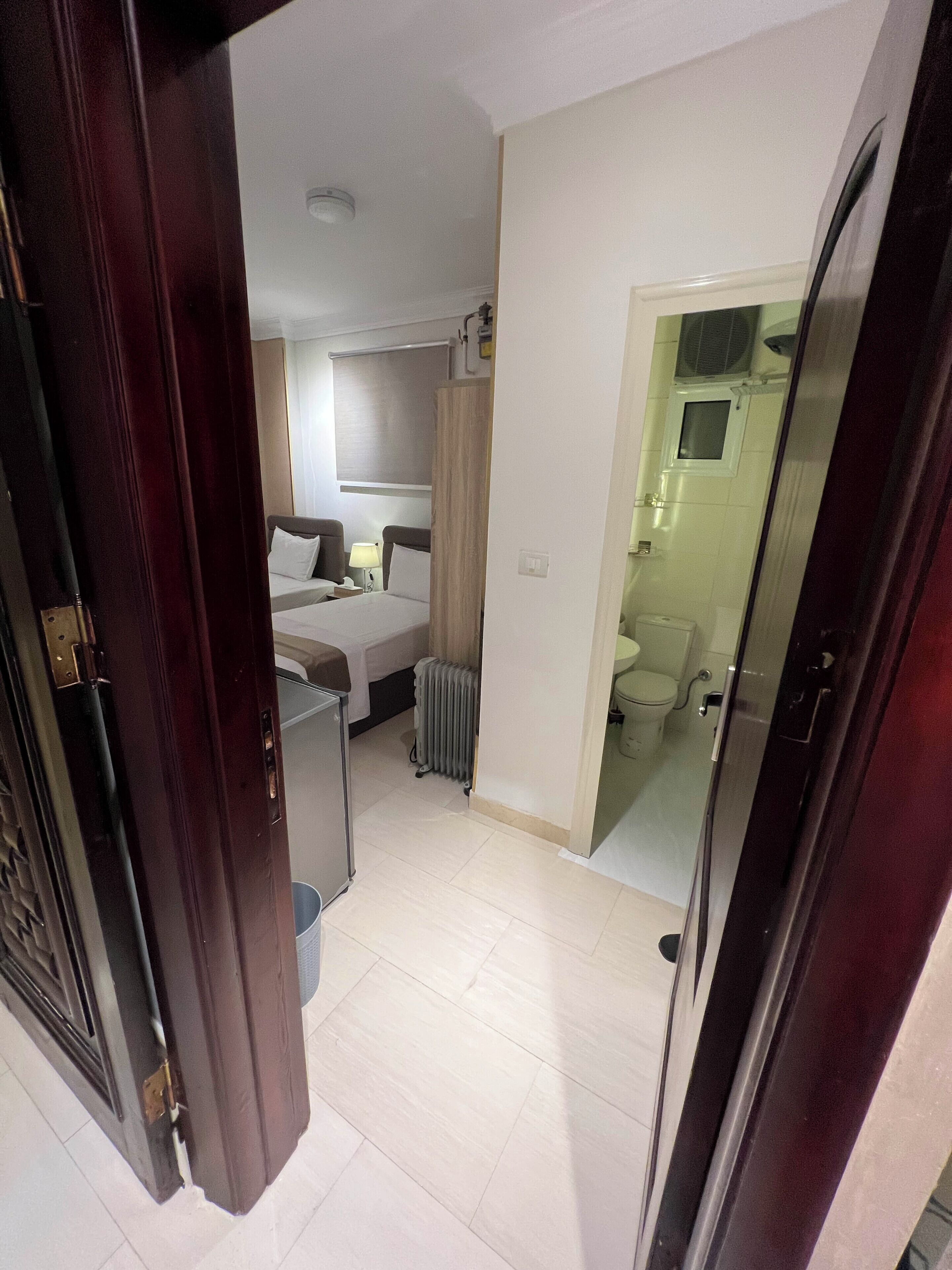Een hotelkamer met een bed, nachtkastje, lamp, radiator en een badkamer zichtbaar door een open deur.