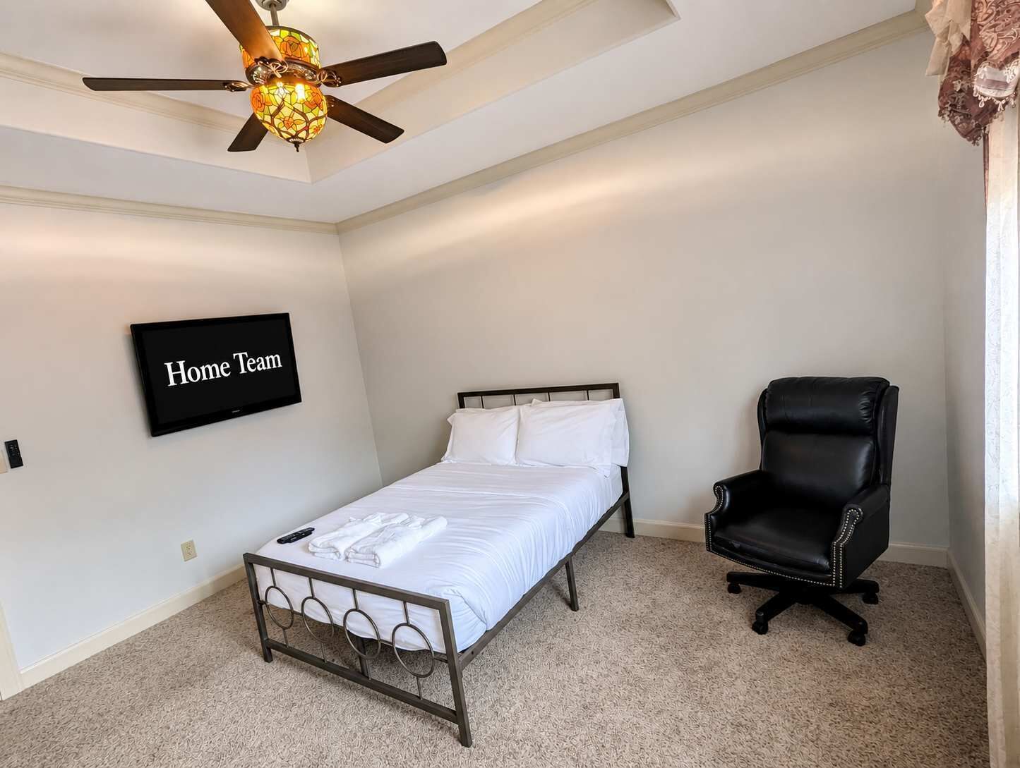 Um quarto com cama, uma cadeira, ventoinha no teto, televisão na parede e piso coberto por carpete.