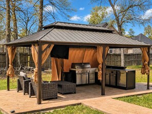 Ett takförsett utomhuskök med grill, inbyggd ugn och en grillplats, omgivet av möbler i rotting och en pergola.
