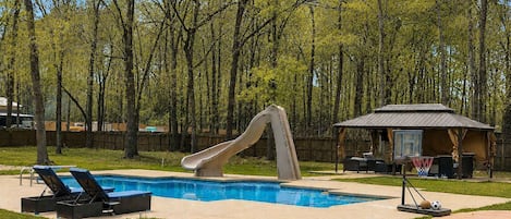 Un jardin avec une piscine, un toboggan, des chaises longues et un petit pavillon.