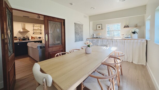 Un grand espace à manger avec une longue table en bois et des chaises en osier, une cuisine en arrière-plan et une fenêtre offrant une vue agréable.