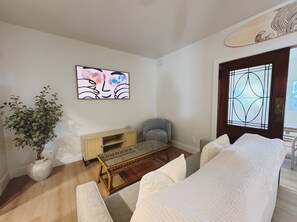 Ein Wohnzimmer mit einer Couch, einem Glastisch, einem grauen Sessel, einem Holzschrank und einer Topfpflanze.