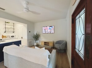 Ein Zimmer mit einem Bett, einem Sessel, einem kleinen Tisch, einer Topfpflanze, einer Mikrowelle und einem Fenster mit Verzierglas.