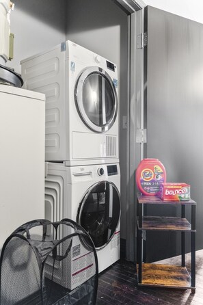 A compact laundry area with a front-loading washer and dryer, a small shelf with laundry detergent, and a mesh laundry basket. - Downtown Dallas Haven | Pool + Theater + Game Room (Dallas)