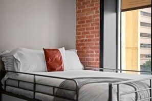 A bed with white bedding and a brown pillow, a metal headboard, a brick wall, and a window with a view of buildings. - Downtown Dallas Haven | Pool + Theater + Game Room (Dallas)