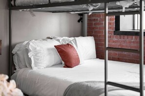 A bunk bed with white bedding and a red pillow, a brick wall, and a window. - Downtown Dallas Haven | Pool + Theater + Game Room (Dallas)
