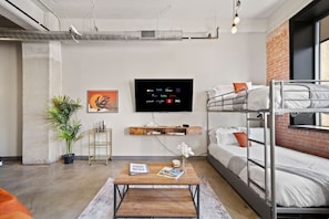 A modern hotel room with bunk beds, a flat-screen TV, a coffee table, and a potted plant. - Downtown Dallas Haven | Pool + Theater + Game Room (Dallas)