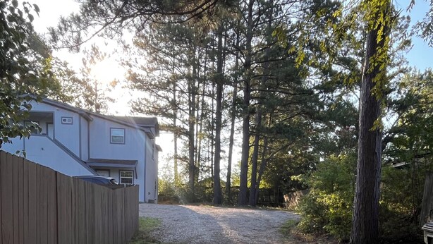 Une allée de gravier mène à une maison entourée d’une clôture en bois, avec des arbres en arrière-plan.