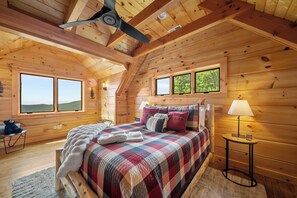 A cozy wooden cabin bedroom with a bed, a nightstand, a lamp, and a window overlooking a forest. - Grandfather Mountain Views, Hot Tub, Game Room & Putting Green (Seven Devils)