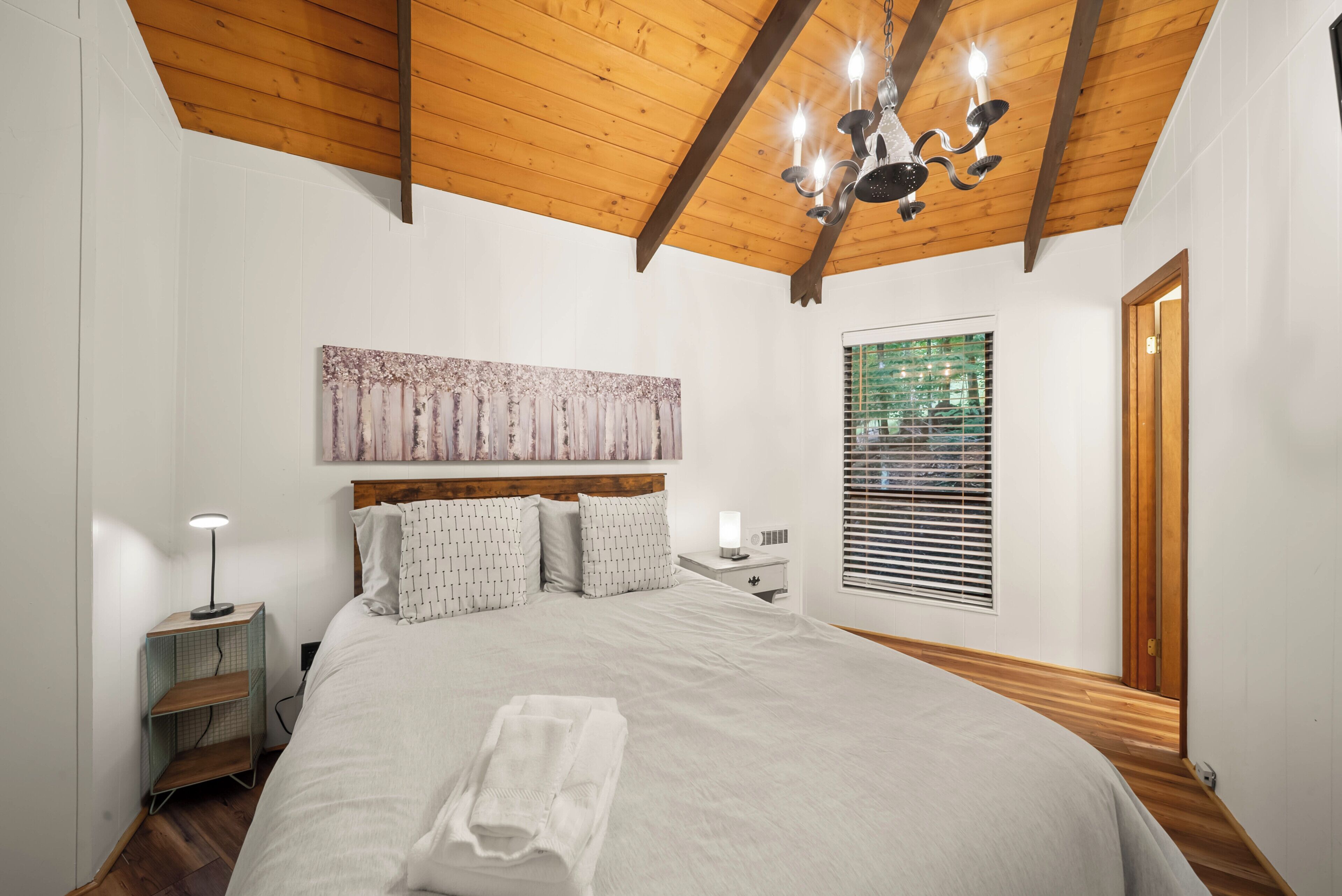Un dormitorio con techo de madera, una cama con cabecera, una mesita de noche, una lámpara, una ventana con persianas y una puerta.