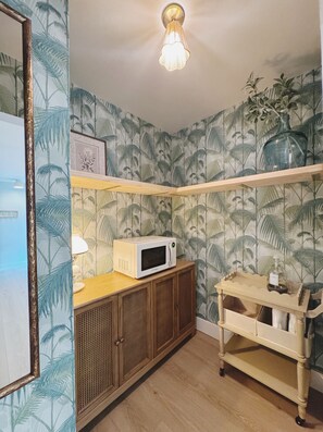 Ein Zimmer mit einer Mikrowelle auf einem Holzkabinent, einem kleinen Tisch mit einer Flasche und einer Wand mit tropischen Blattmotiven.