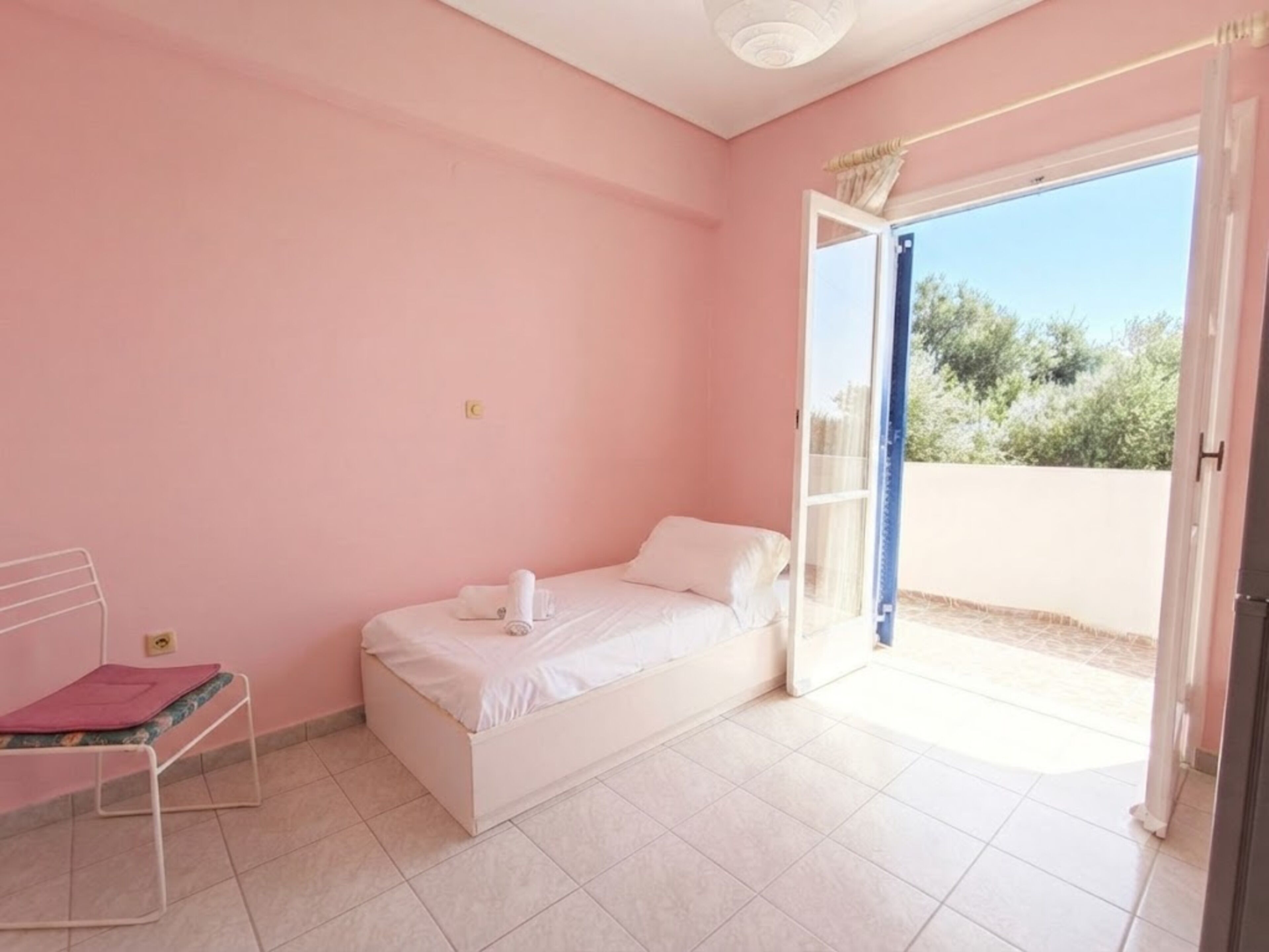 Une chambre avec un lit simple, une petite table et une fenêtre avec une porte ouverte donnant sur l’extérieur.