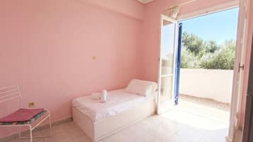 Une chambre avec un lit simple, une petite table et une fenêtre avec une porte ouverte donnant sur l’extérieur.