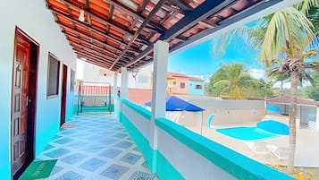 Um corredor externo coberto com piso de azulejos, que dá acesso a uma área de piscina com guarda-sol azul.