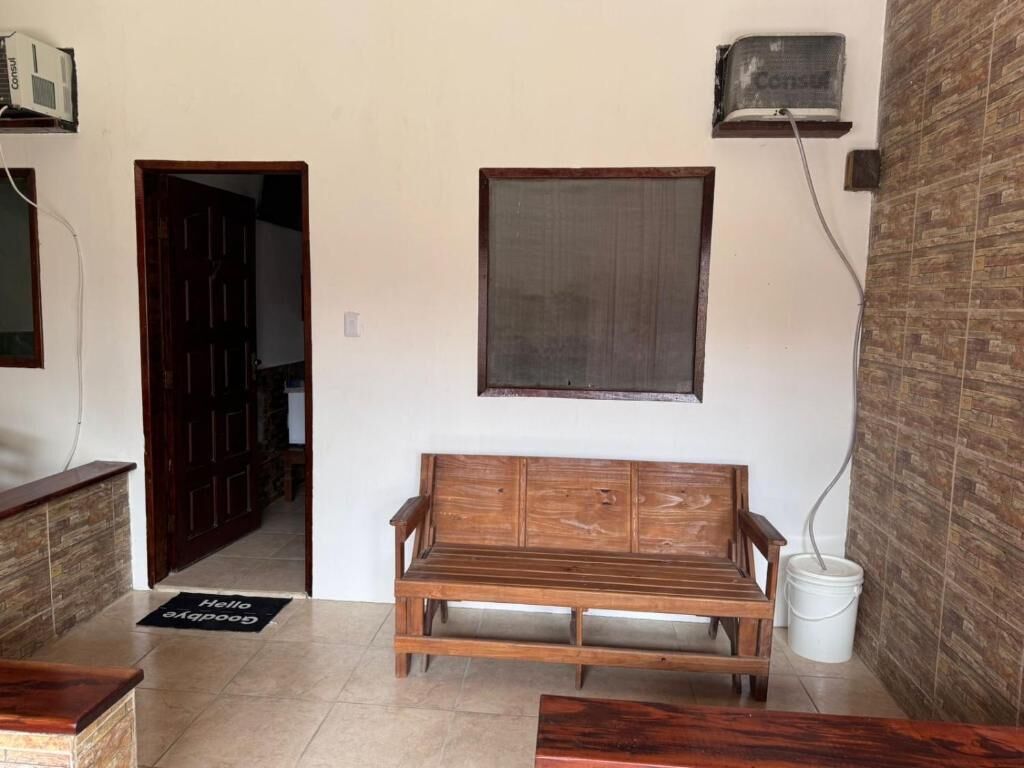 Una habitación con un banco de madera, una puerta, un aire acondicionado instalado en la pared y un balde blanco.