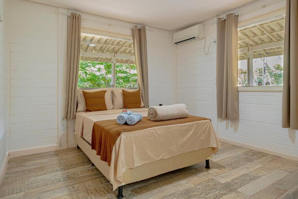 Una cama individual con una colcha color beige, dos toallas enrolladas y un plaid marrón en una habitación con paredes blancas y suelo de madera.
