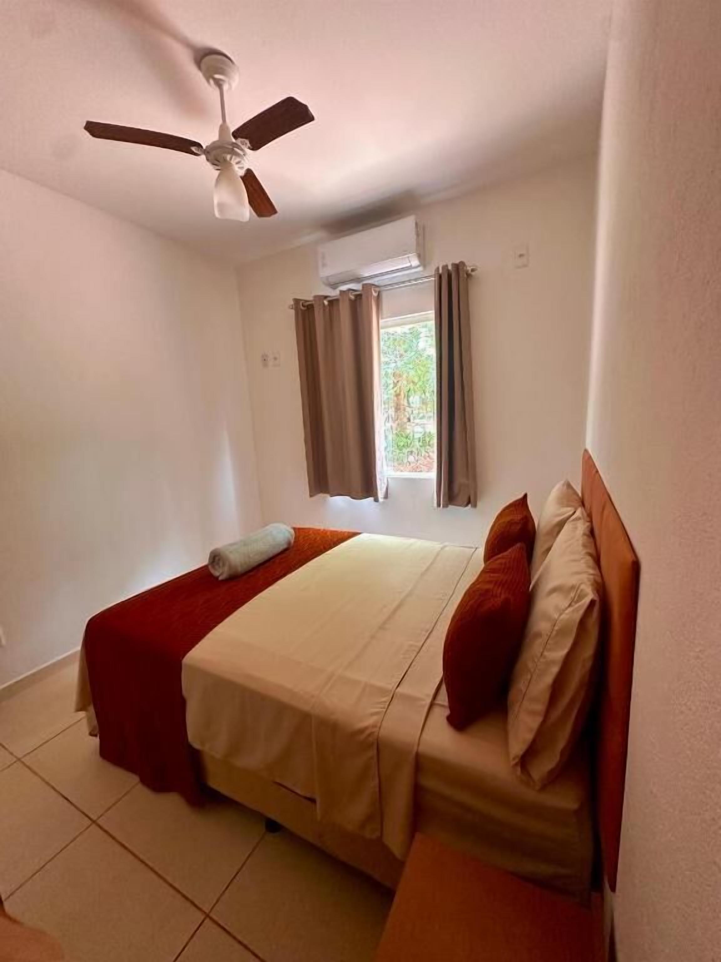 A hotel room with a bed, a ceiling fan, a window with curtains, and a view of greenery outside.
