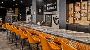 Zona de bar con una barra alargada, varios taburetes y una pared decorada con logotipos de equipos deportivos.