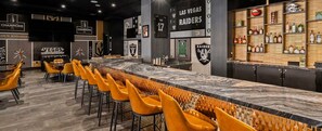 A bar area with a long counter, multiple stools, and a wall adorned with sports team logos.