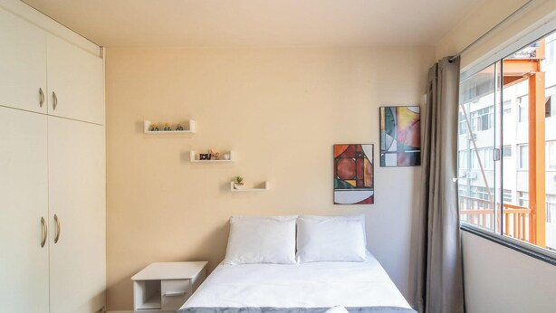 A hotel room with a bed, a small bedside table, a wardrobe, and a window with a view of buildings. - Apart w/ Side sea View, 50m From the Beach Mrd0061 (Balneário Camboriú)