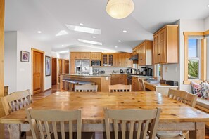 A modern kitchen with wooden cabinets, a central island, and a dining area with a large wooden table and chairs. - Miwok Escape, by Vinifera Homes (Bodega Bay)