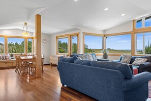 A spacious living area with a large blue sofa, wooden dining table, and multiple windows offering views of the outdoors. - Miwok Escape, by Vinifera Homes (Bodega Bay)