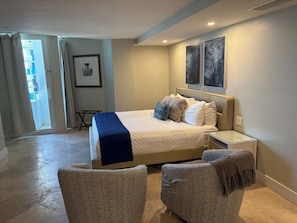 Ein Schlafzimmer mit einem Bett, zwei Nachttischen, einem Sessel, einem kleinen Tisch und einer Tür, die auf einen Balkon führt.