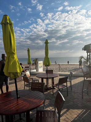 A beachside patio with wooden tables and chairs, green umbrellas, and a view of the ocean. - Oceanfront Sunrise Escape Condo | Beach and Pool (Fort Lauderdale)