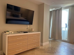 A room with a flat-screen TV mounted on the wall, a wooden cabinet with drawers, and a sliding glass door leading to an outdoor area. - Oceanfront Sunrise Escape Condo | Beach and Pool (Fort Lauderdale)