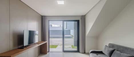 A modern living room with a flat-screen TV on a wooden console, a grey sofa, and a sliding glass door leading to an outdoor area.